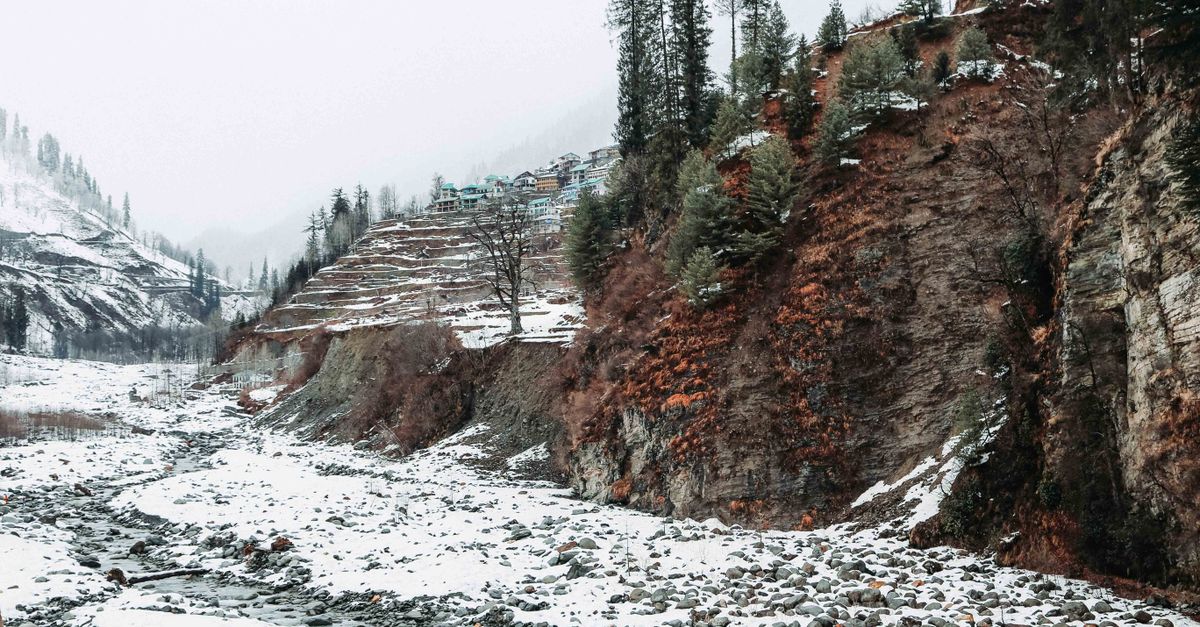Picturesque view of uneven mounts with growing trees near shabby terrain covered with snow and small stones under serene sky in winter